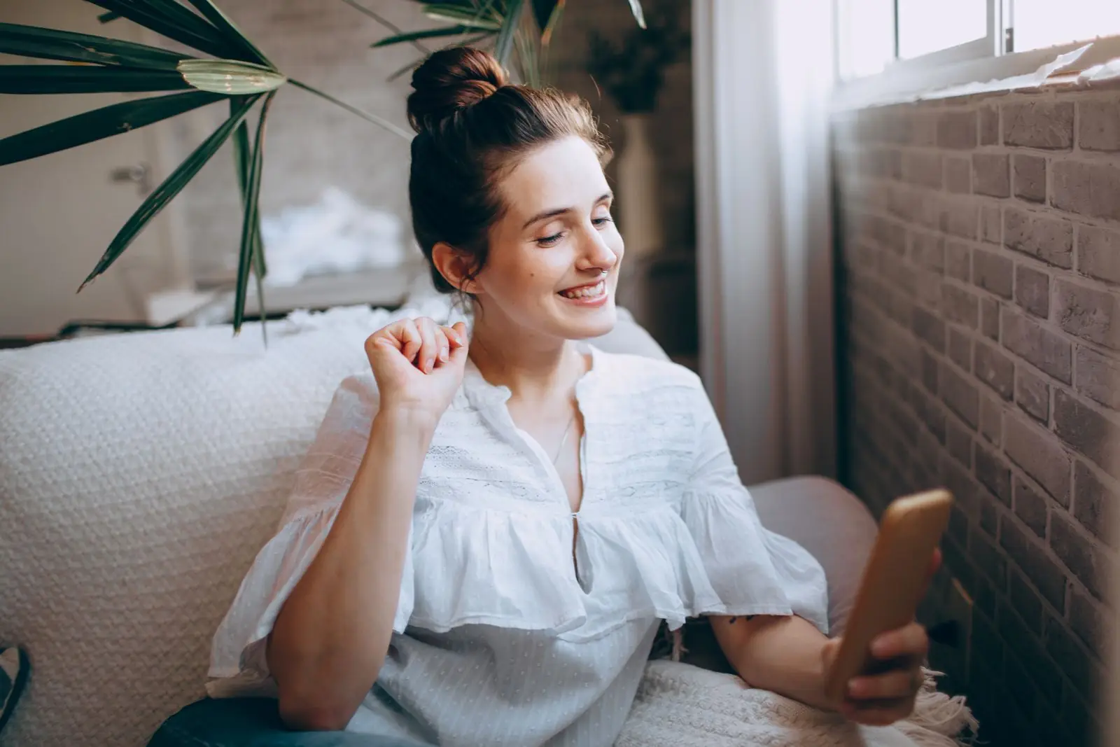 A woman smiles at her phone while sitting on a sofa at home.