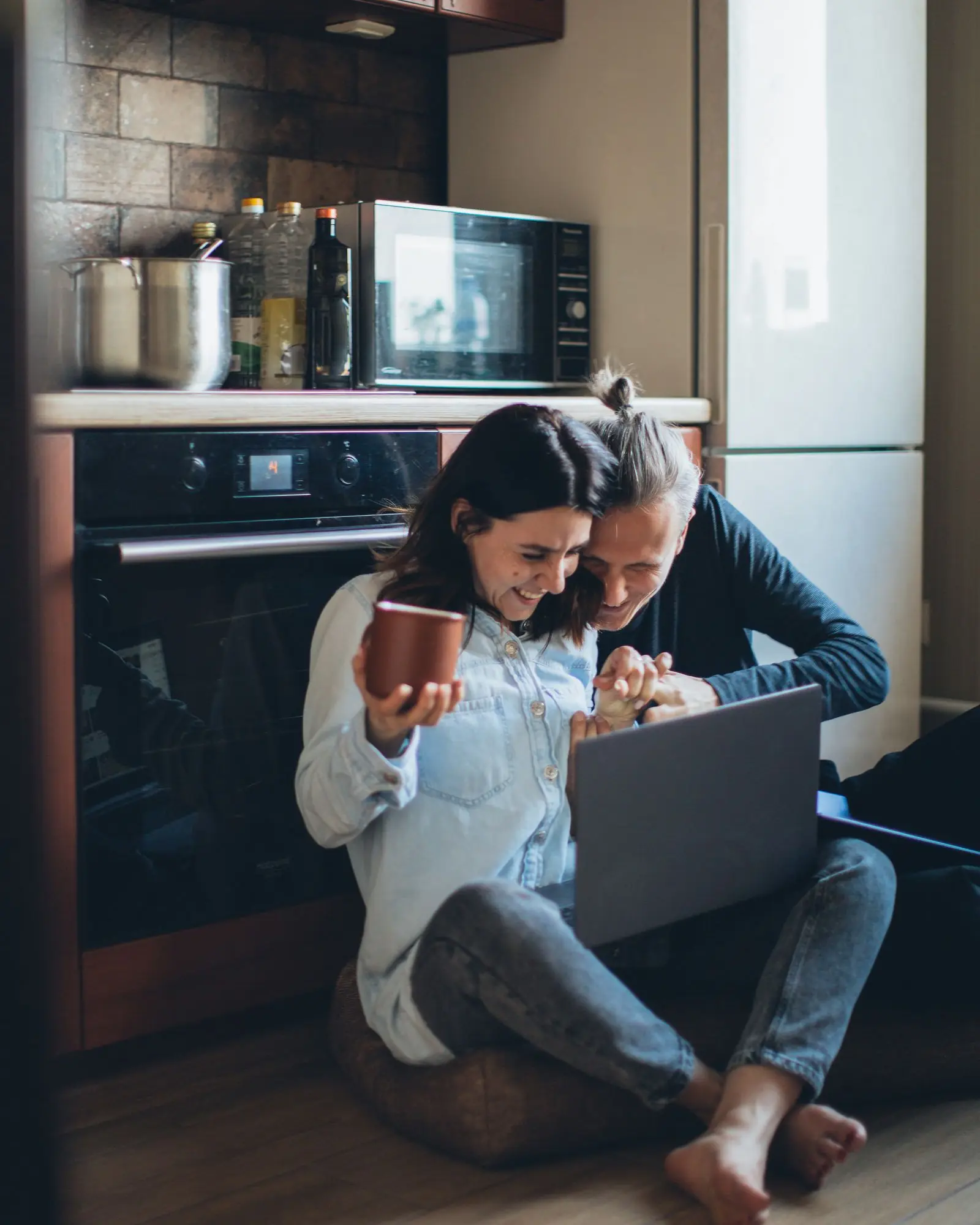 Two people sit on a kitchen floor, smiling at a laptop together.
