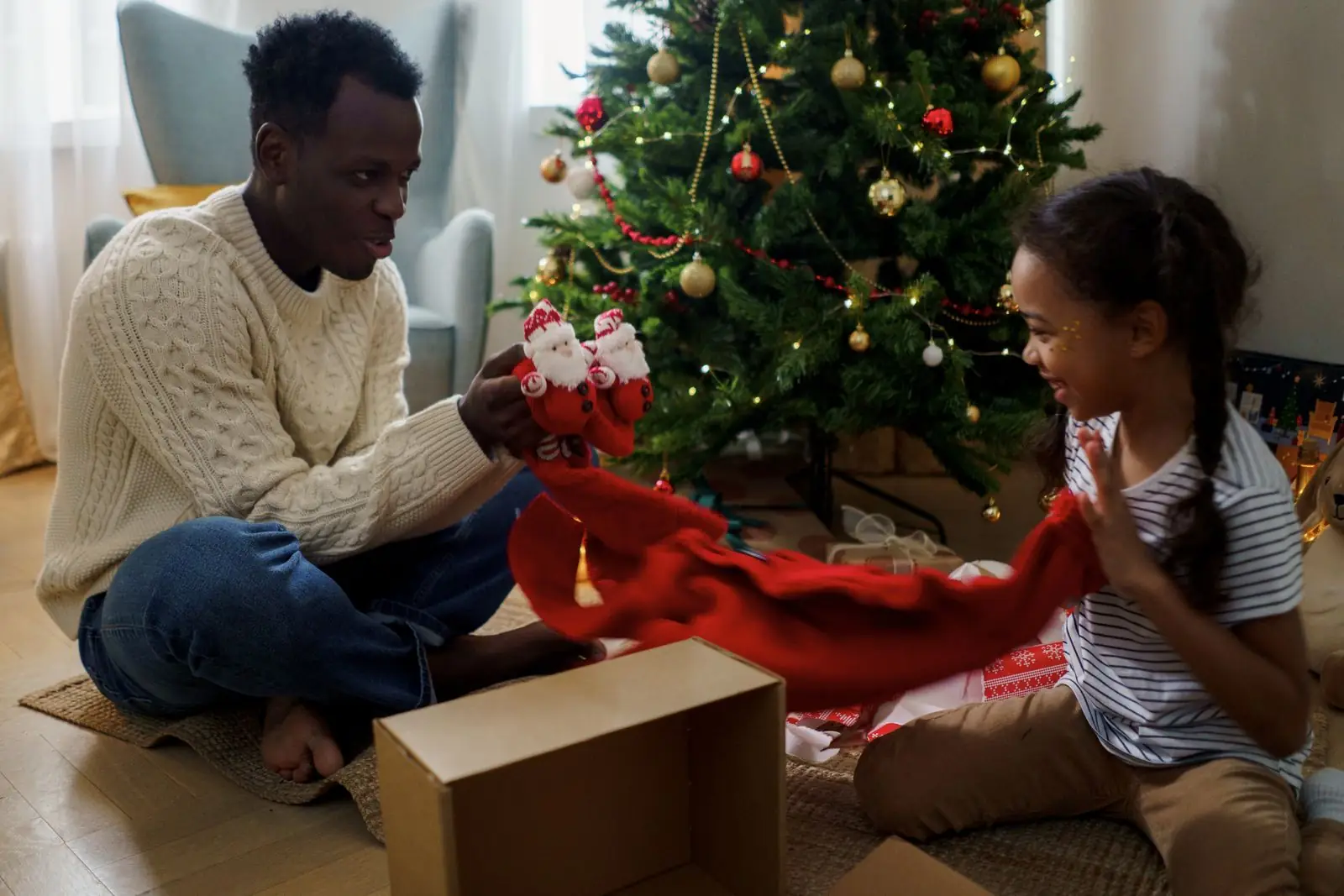 A father and daughter open a present together by a Christmas tree.